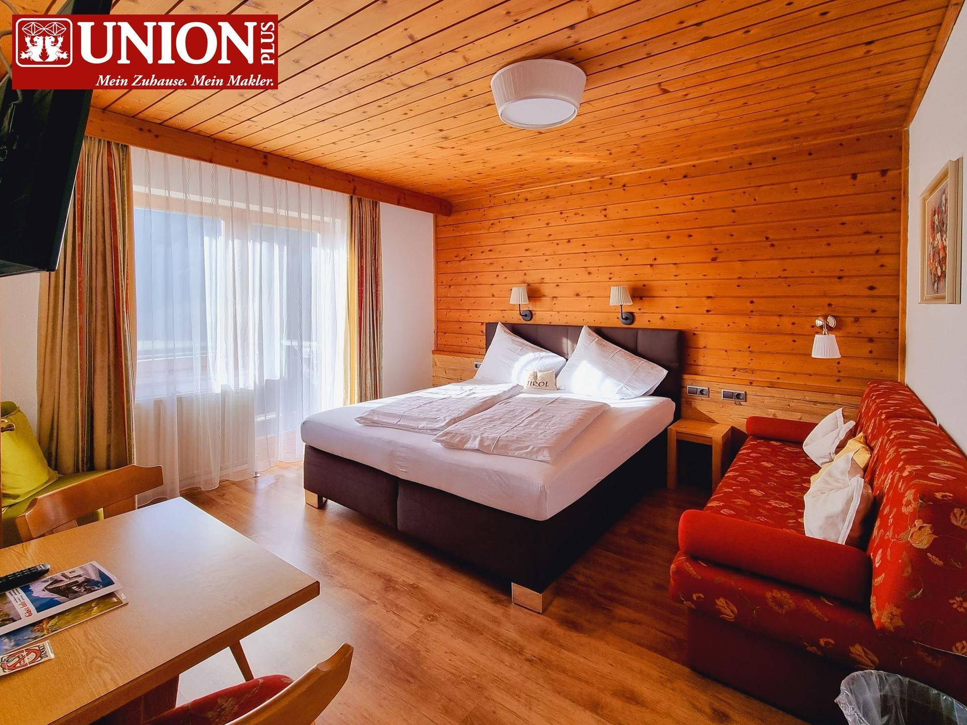 Gemütliches Schlafzimmer mit Holzwänden, Doppelbett und rotem Sofa, hell durch großes Fenster.
