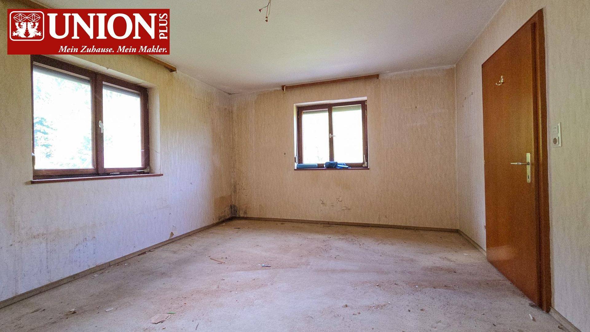 Leerer Raum mit zwei Fenstern und einer Holztür, bereit für Renovierungsarbeiten.