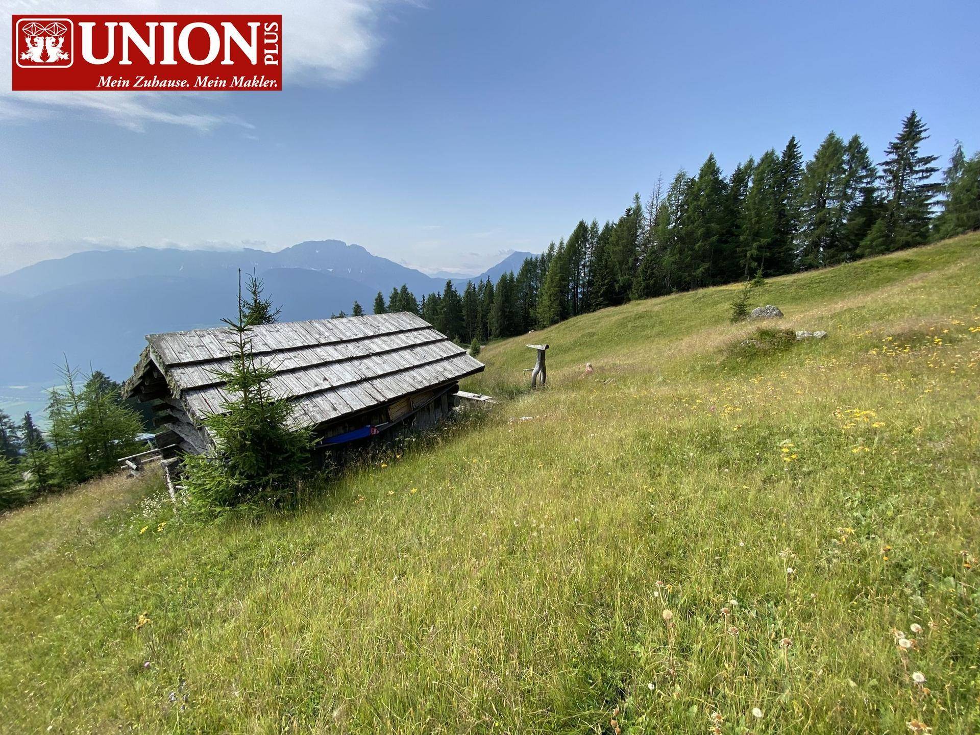 Rustikale Holzhütte auf einer sonnigen Bergwiese mit weitem Blick auf die umliegende Alpenlandschaft.