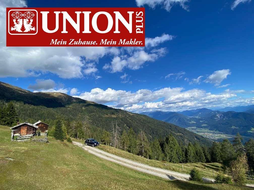 Panoramablick über eine Berglandschaft mit Schotterweg, Holzhütten und einem Fahrzeug unter blauem Himmel.
