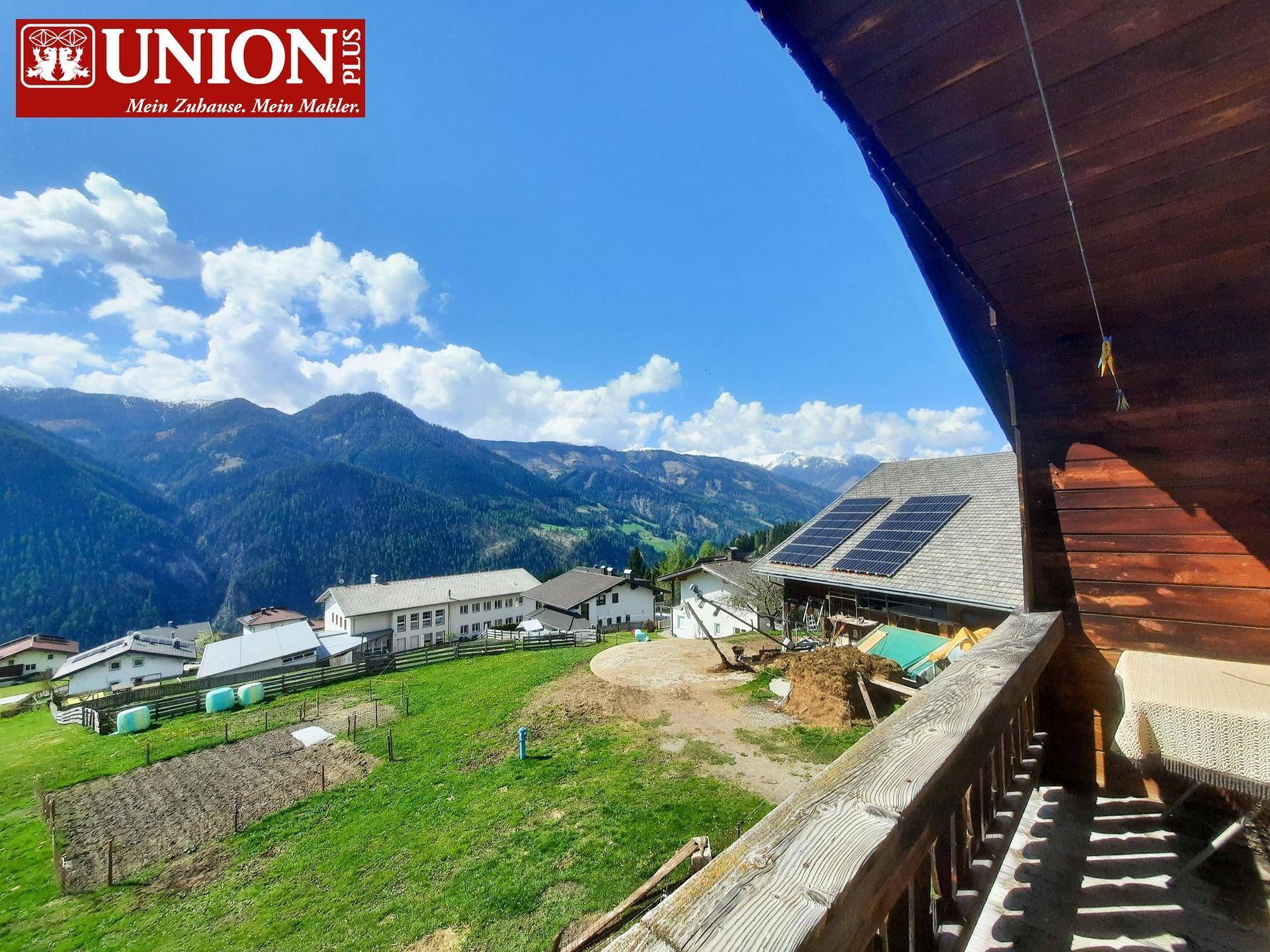Blick von einem Holzbalkon auf eine weite Berglandschaft mit weiteren Häusern und einer Photovoltaikanlage.