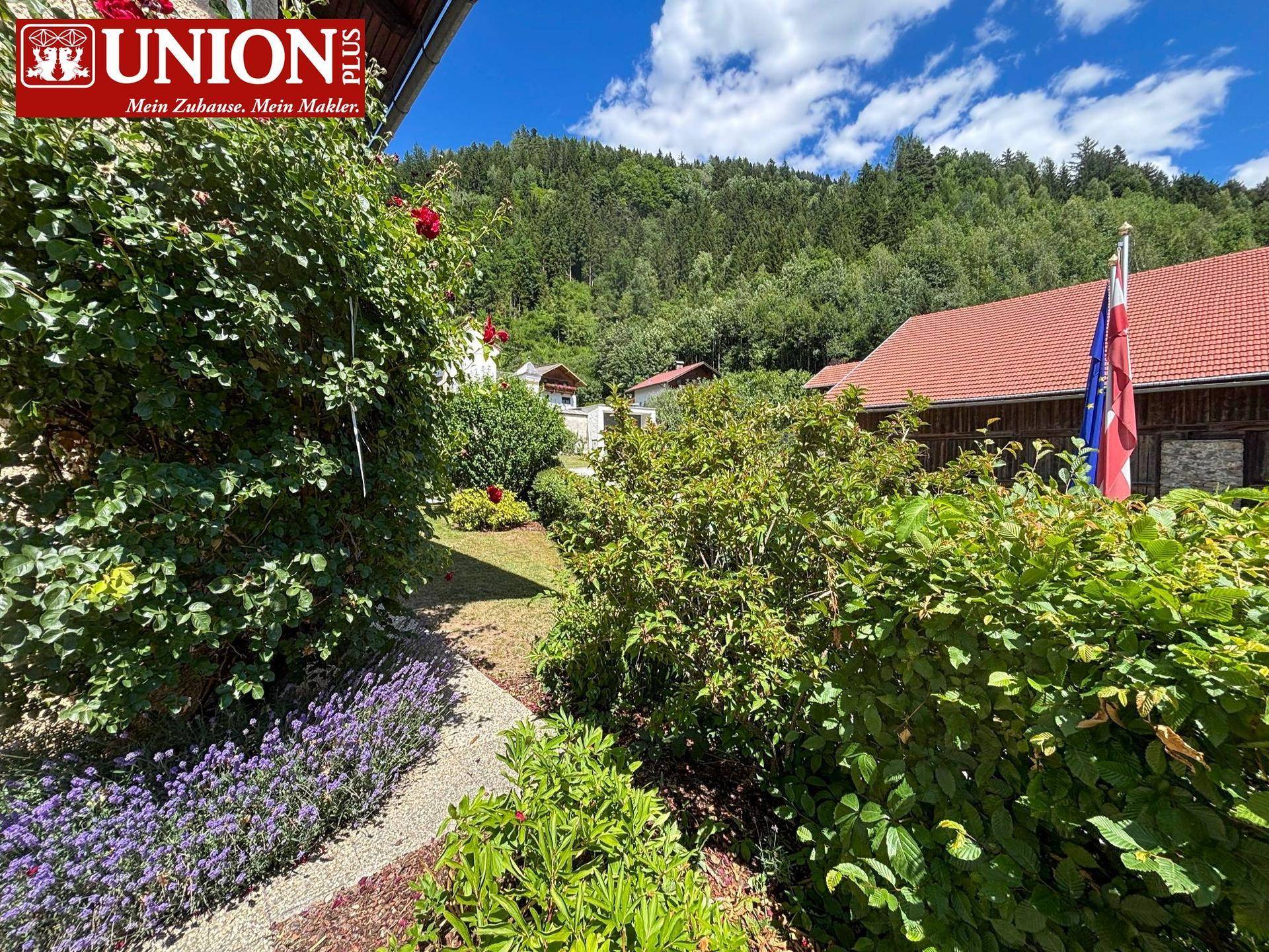 Idyllischer Gartenweg umgeben von üppigem Grün, Rosen und Lavendel mit Blick auf bewaldete Hügel.