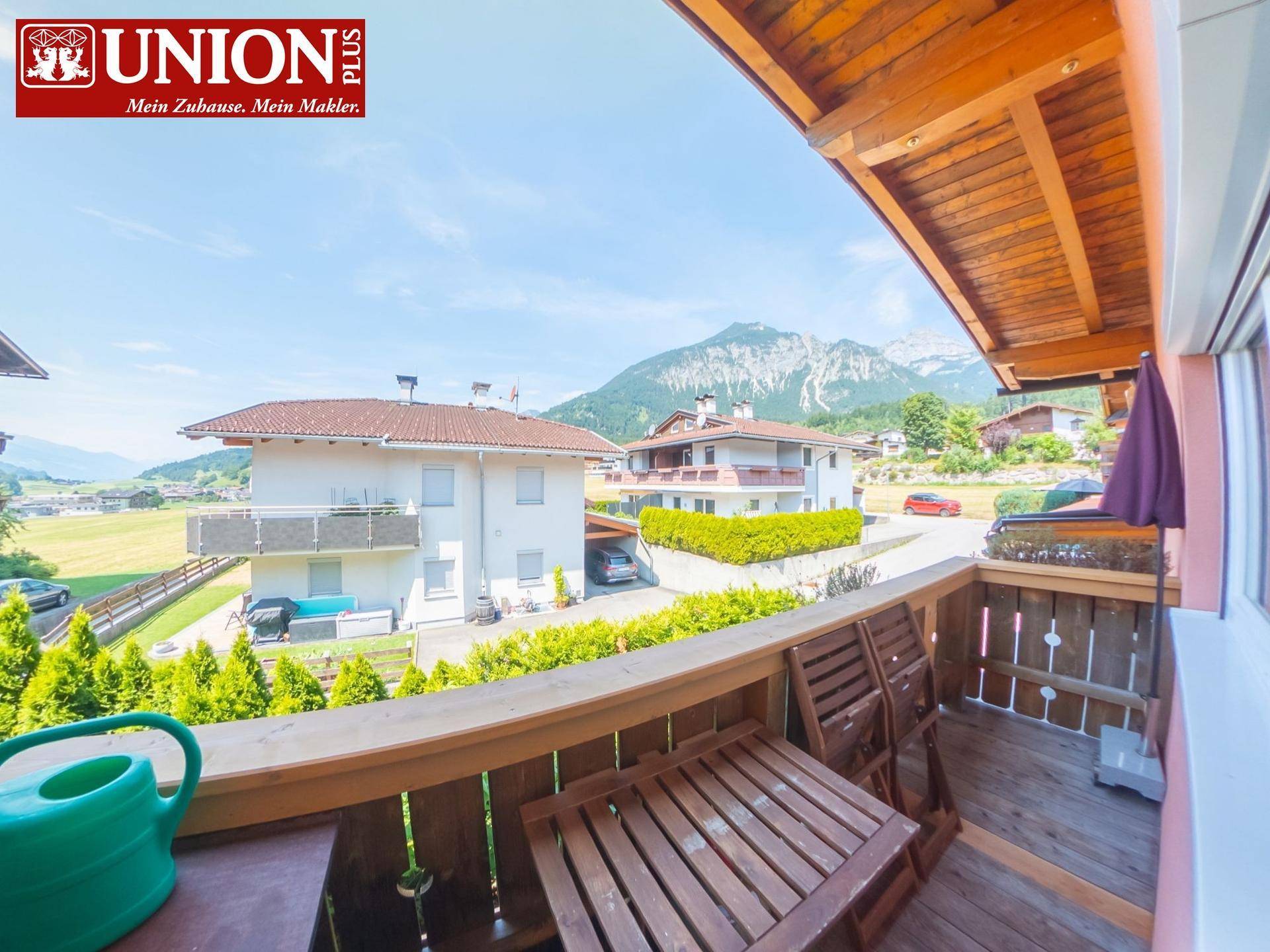 Holzbalkon mit Sitzgelegenheiten und weitem Blick auf die Berge und umliegende Häuser.