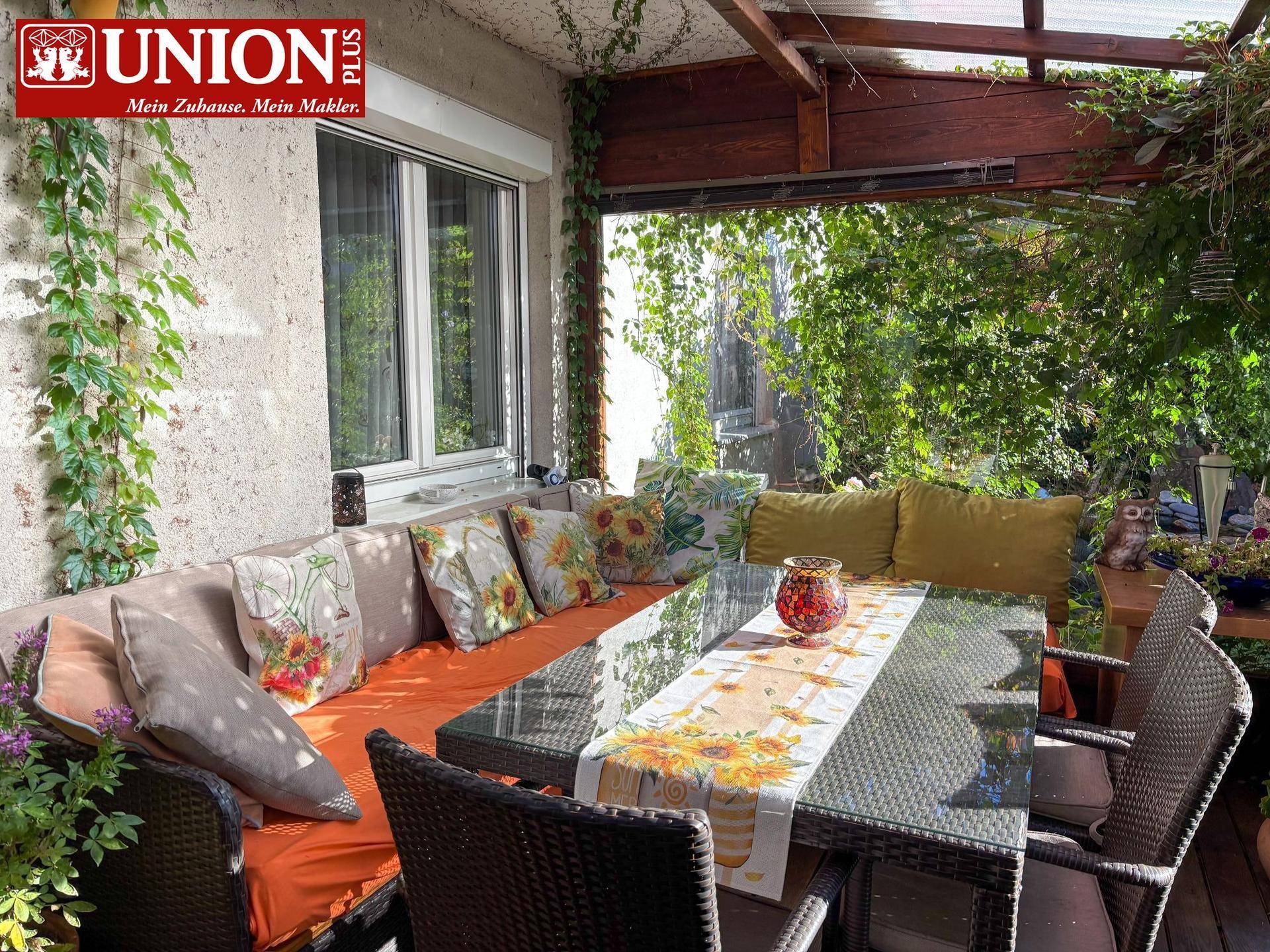 Überdachte Terrasse mit gemütlicher Sitzgruppe, Esstisch und üppiger Begrünung.