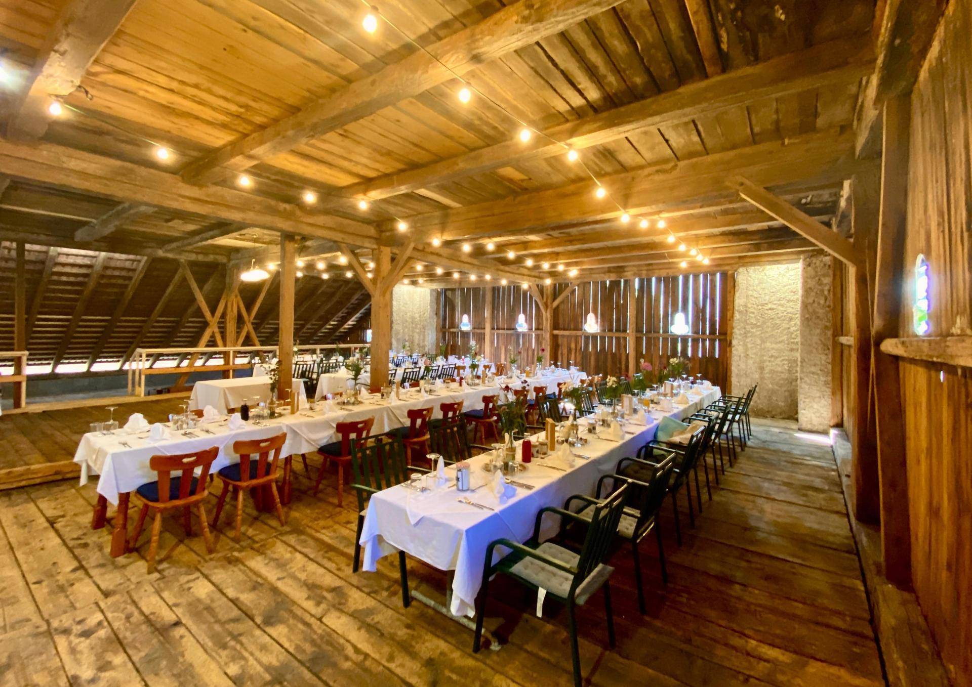 Großer, rustikaler Festsaal mit langen Tischen, Holzbalken und warmen Lichterketten für eine gemütliche Atmosphäre.
