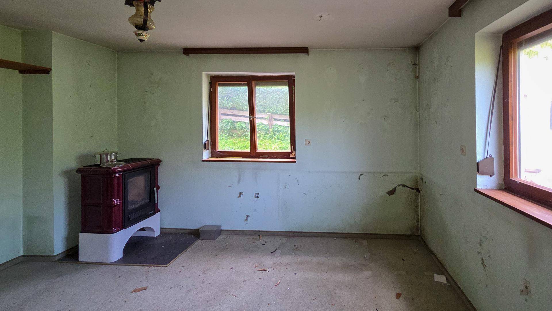 Leeres Zimmer mit hellgrünen Wänden, einem traditionellen Kachelofen und Holzfenstern.
