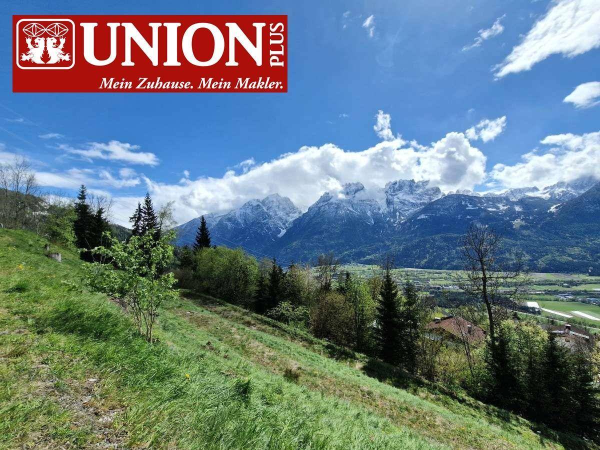 Weitläufiger Blick auf die Alpen mit grünen Hügeln und vereinzelten Häusern an einem sonnigen Tag.