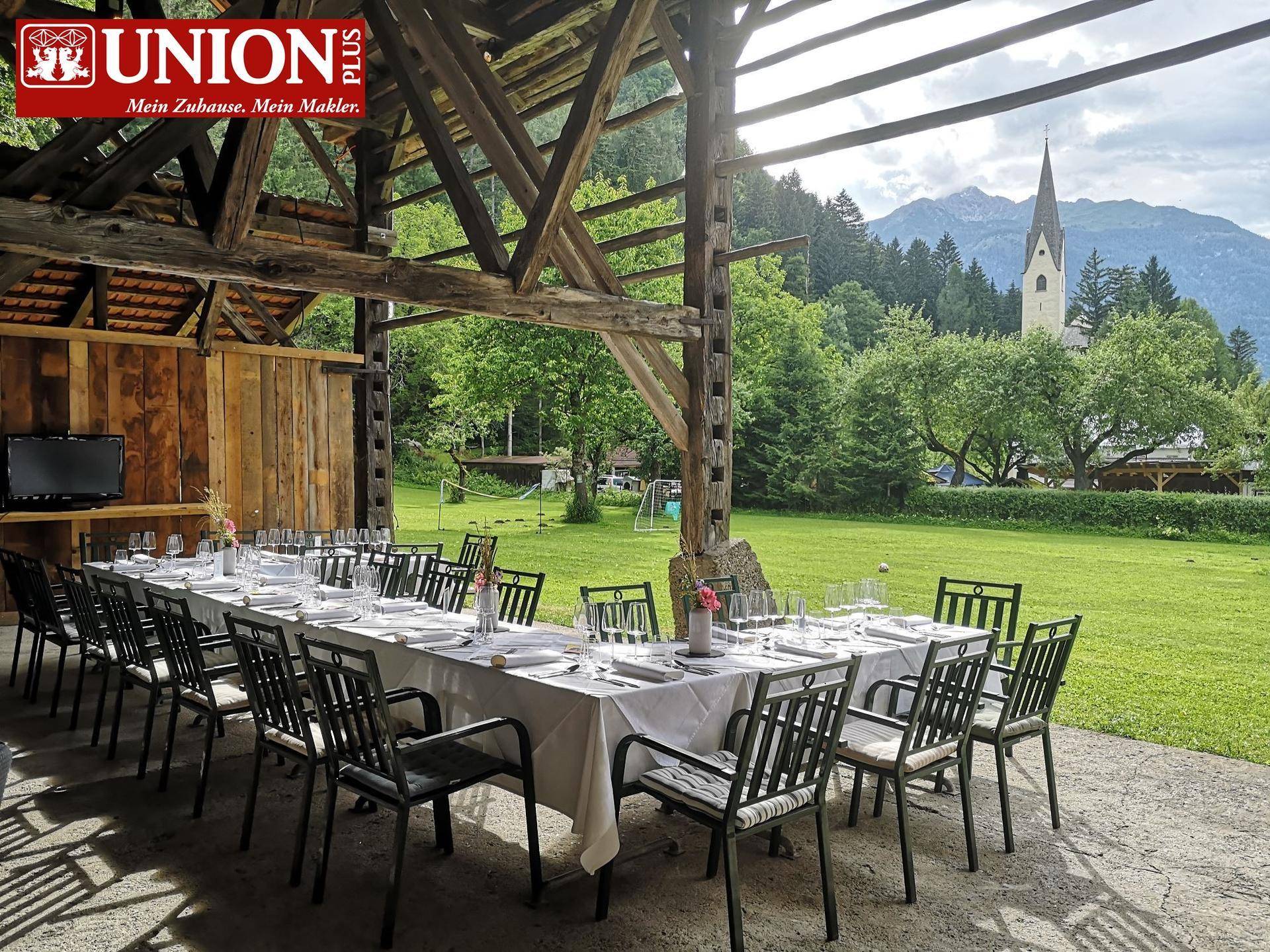 Großer überdachter Essbereich im Freien mit langen Tischen und Stühlen, umgeben von grüner Natur und Bergblick.