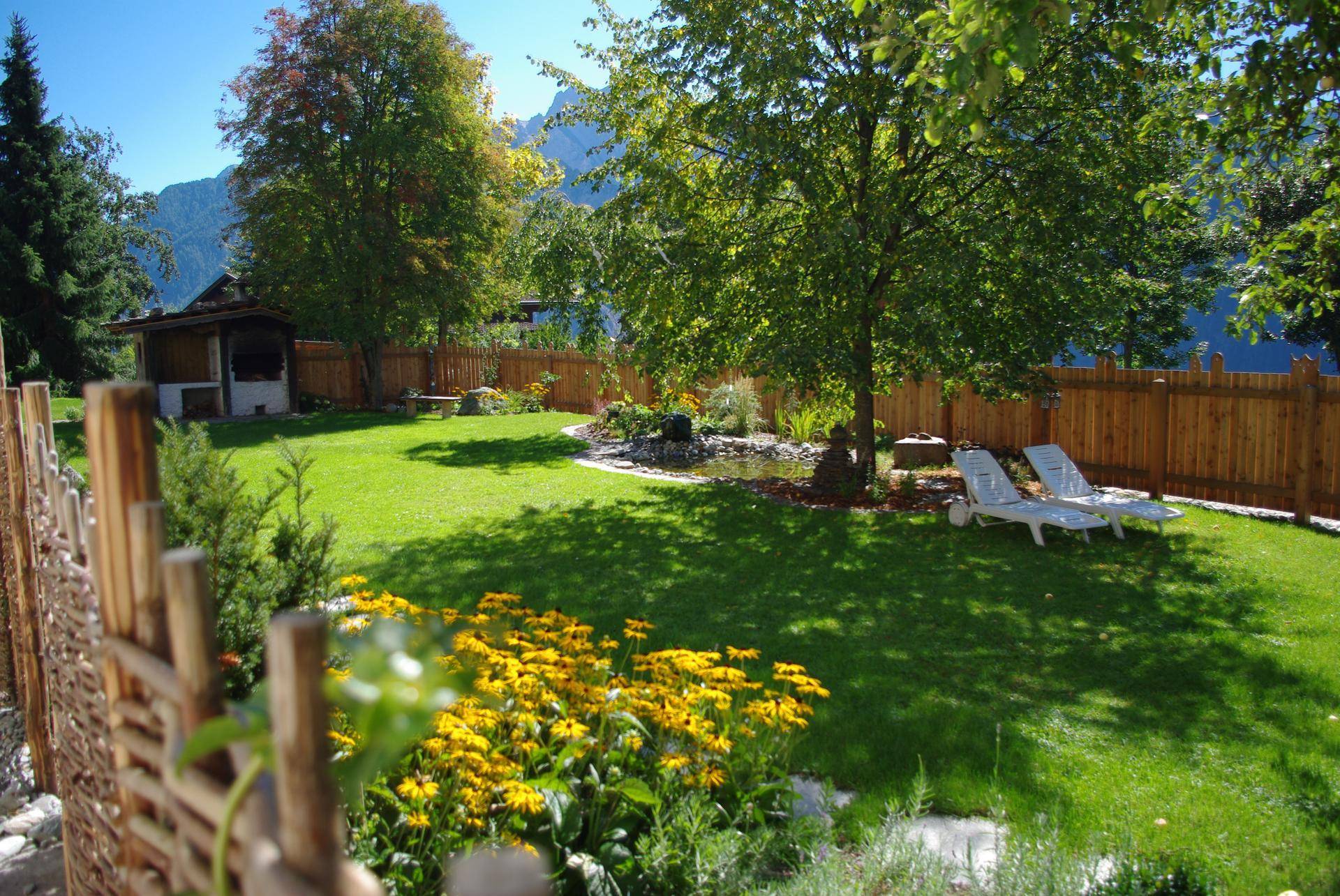 Idyllischer Gartenbereich mit Rasenfläche, Blumenbeeten, einem kleinen Teich und Liegestühlen.