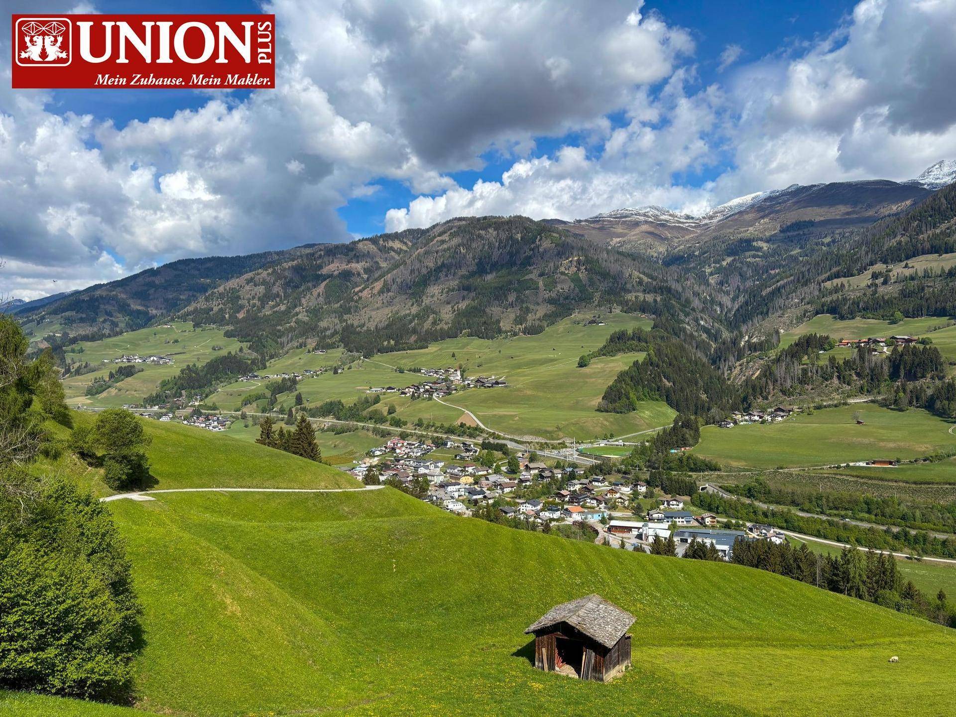 Idyllische Hügellandschaft mit einem kleinen Dorf und schneebedeckten Berggipfeln im Hintergrund.