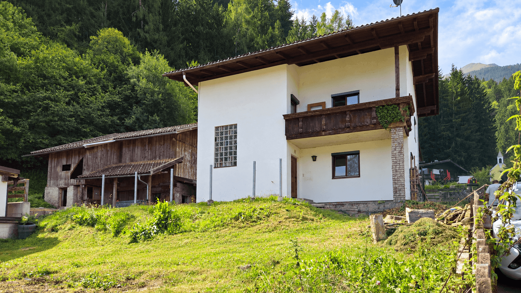 Weißes Wohnhaus mit traditionellem Holzbalkon und angrenzendem Scheunengebäude.