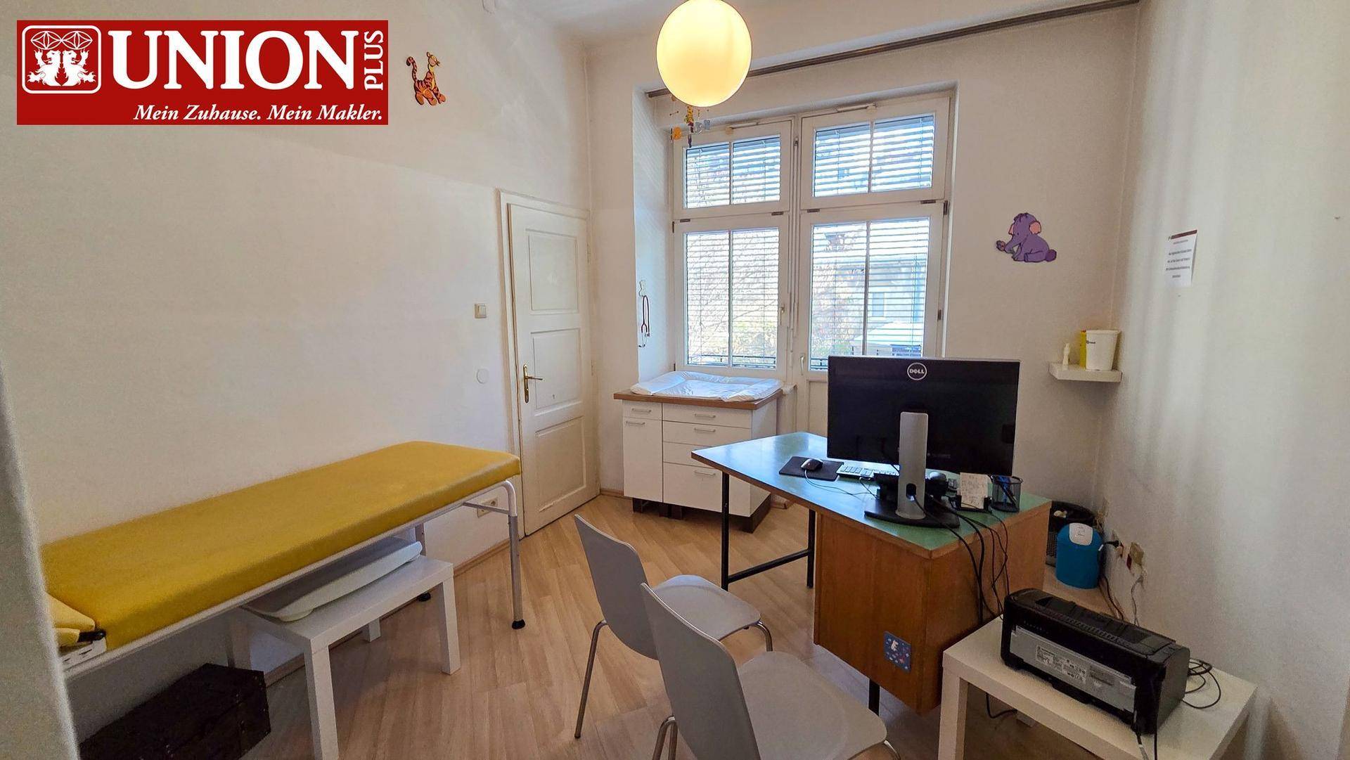 Heller Behandlungsraum mit gelber Untersuchungsliege, Schreibtisch und großem Fenster.