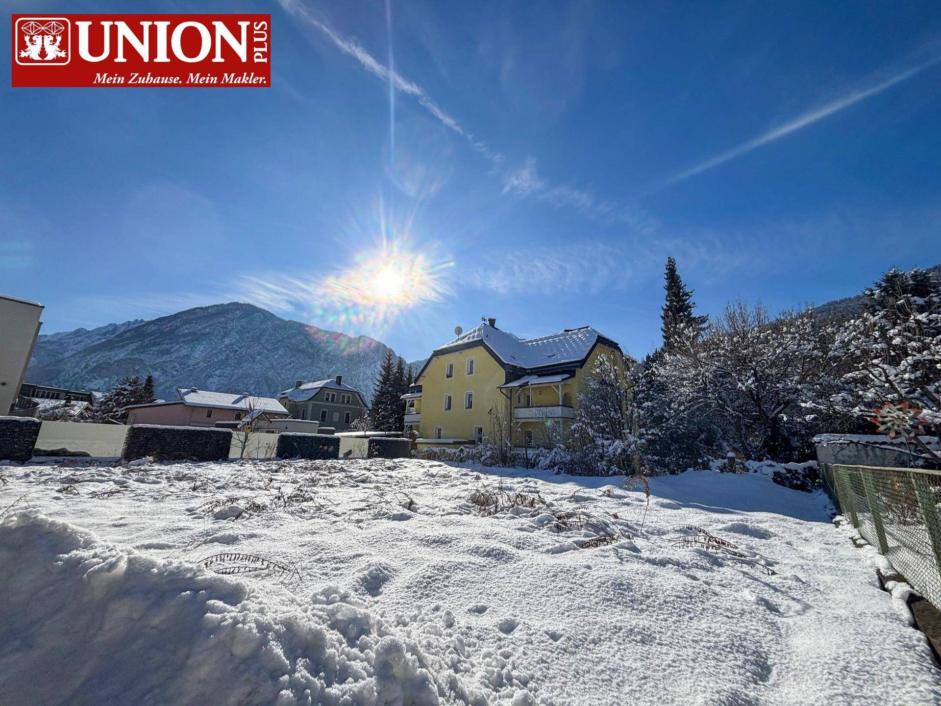 Winterlandschaft mit einem gelben Haus, umgeben von Schnee und Bergen unter einem strahlend blauen Himmel.