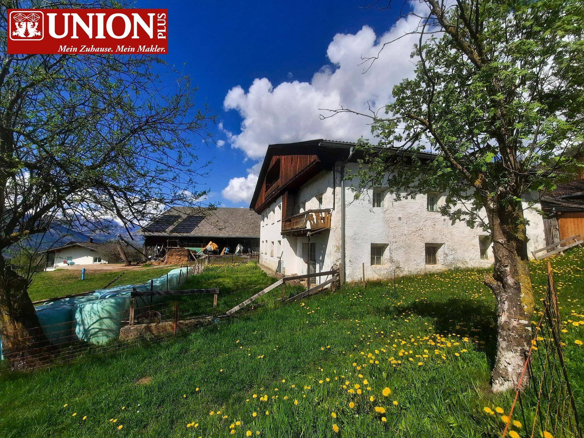 Ländliches Anwesen mit traditionellem Haus, umgeben von grünen Wiesen, Bäumen und einem weiteren Gebäude mit Solaranlage.
