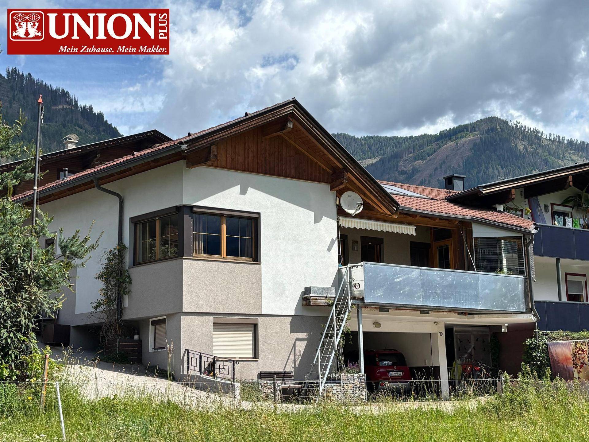 Charmantes Haus mit traditioneller Architektur, Balkon und Carport, umgeben von einer malerischen Berglandschaft.