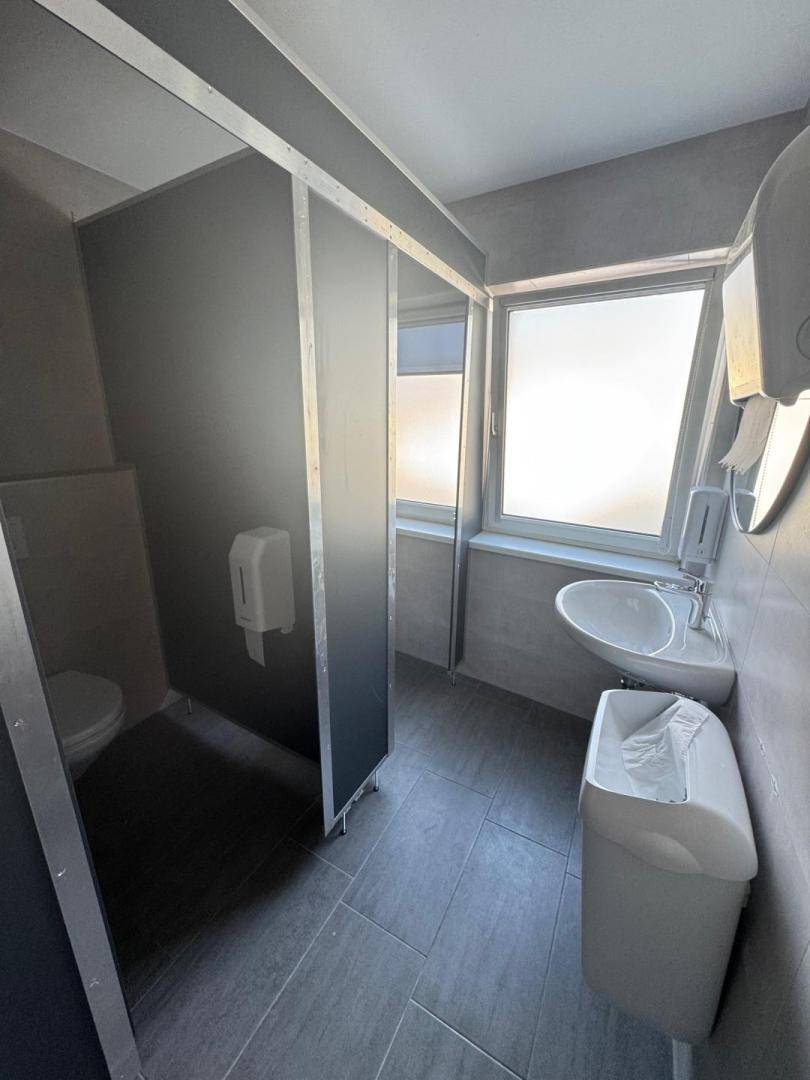 Moderner Toilettenraum mit grauen Fliesen, mehreren Waschbecken und Toilettenkabinen.