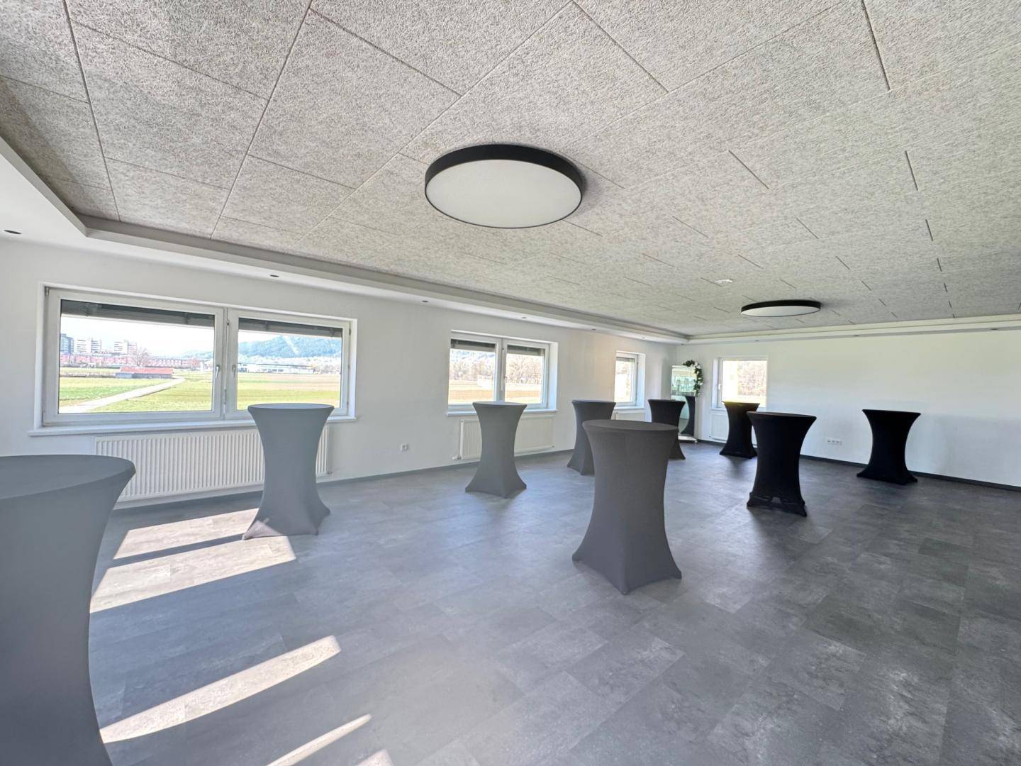Heller Konferenzraum mit grauen Stehtischen, großen Fenstern und Blick auf die umliegende Natur.