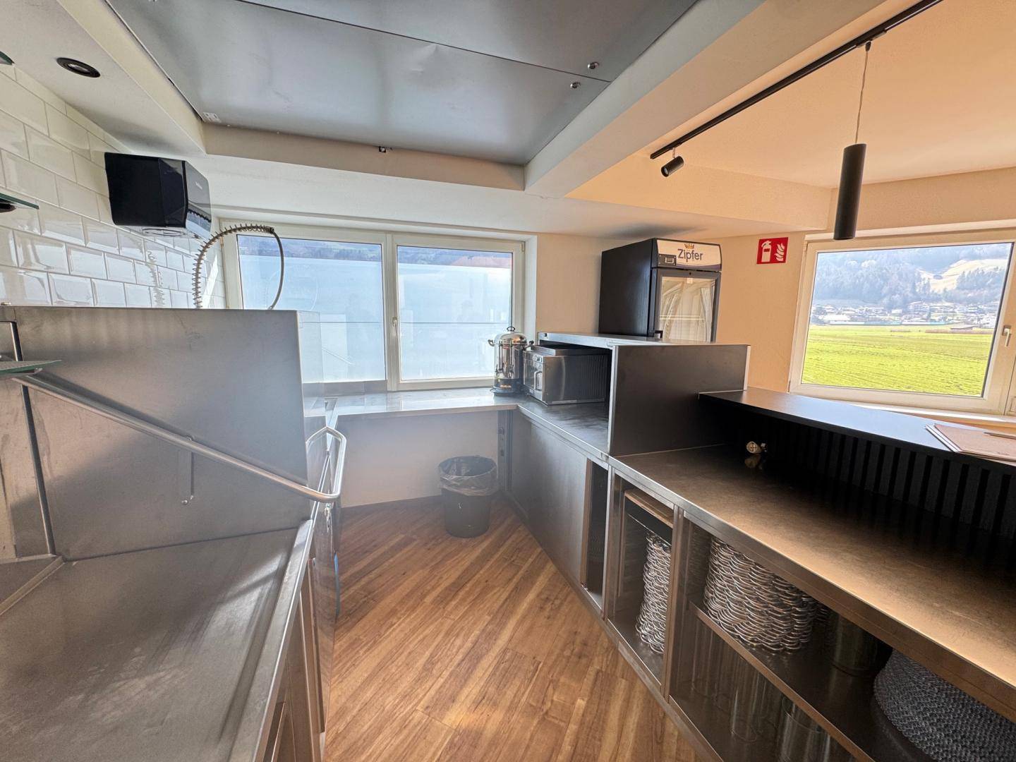 Funktionale Küchenzeile mit Edelstahlflächen, kleinem Kühlschrank und Fenstern mit Blick ins Grüne.