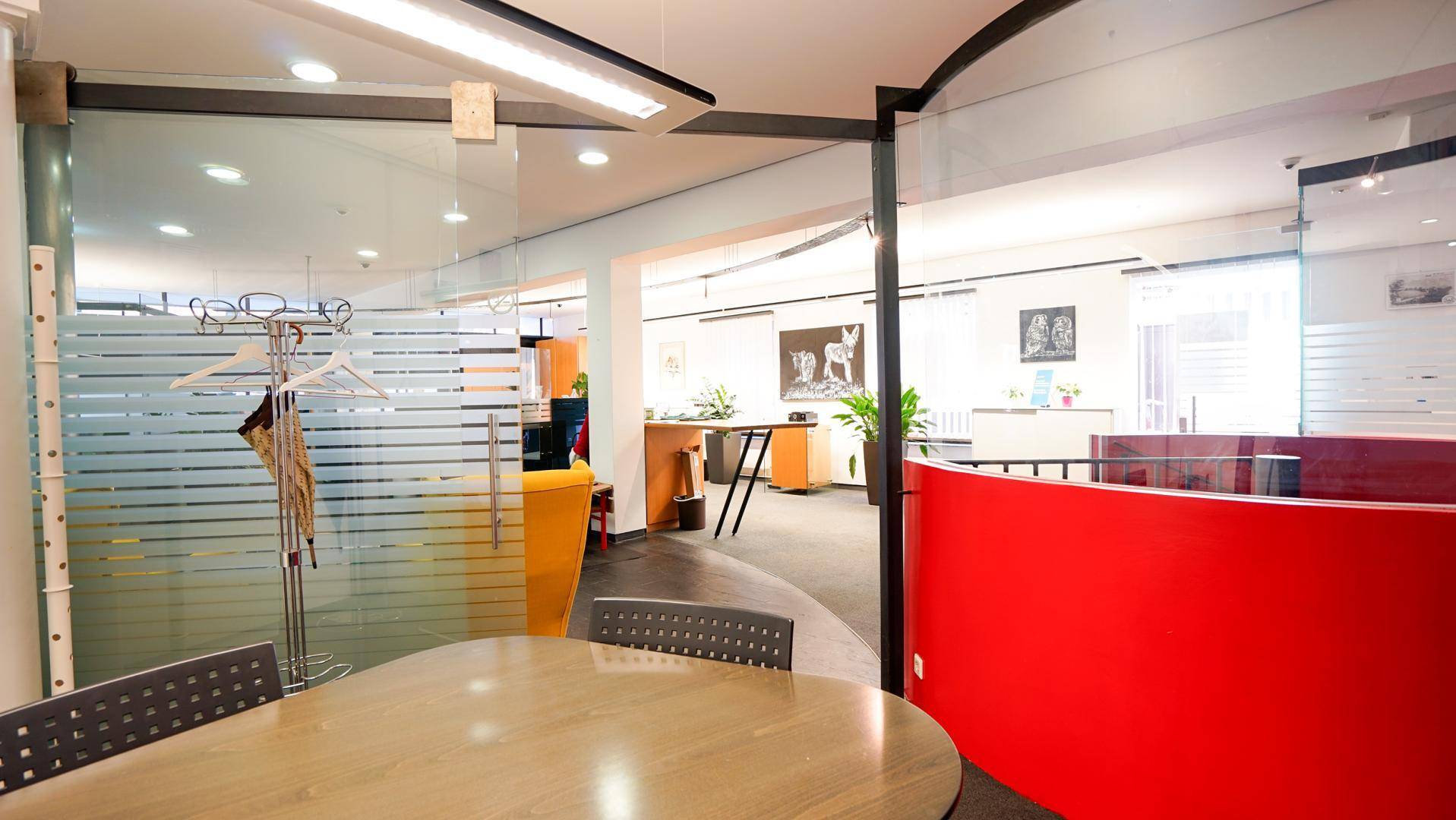 Büro-Besprechungsraum mit Holztisch, Bürostühlen und Blick auf den Empfangsbereich mit roten Akzenten.