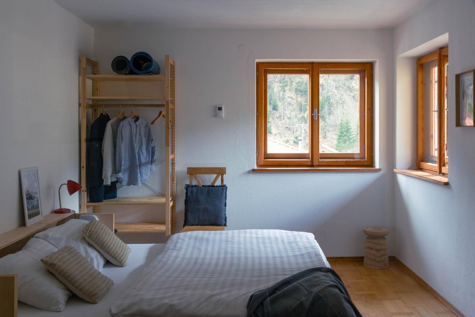 Helles Schlafzimmer mit Holzboden, offenem Kleiderschrank und zwei Fenstern für Tageslicht.