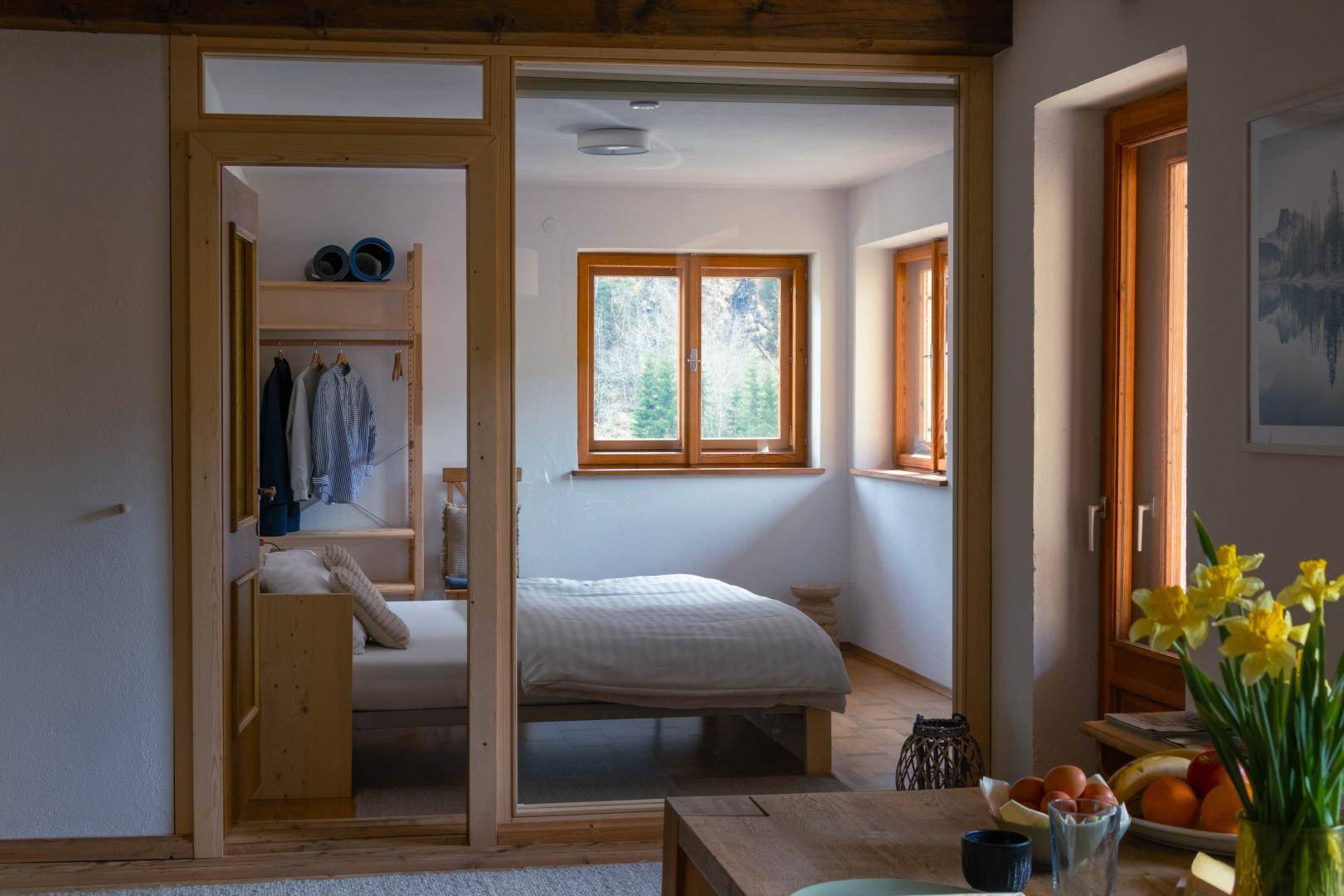 Heller Wohnbereich mit Holztisch und Blick durch eine Glaswand in ein Schlafzimmer.