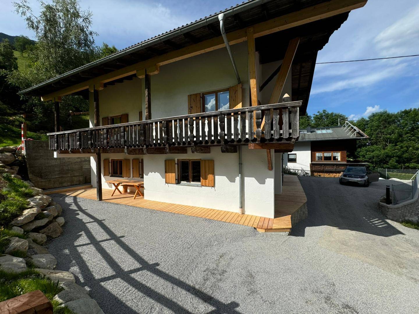 Alpines Haus mit Holzbalkon, Terrasse und Kiesauffahrt, eingebettet in die Berglandschaft.