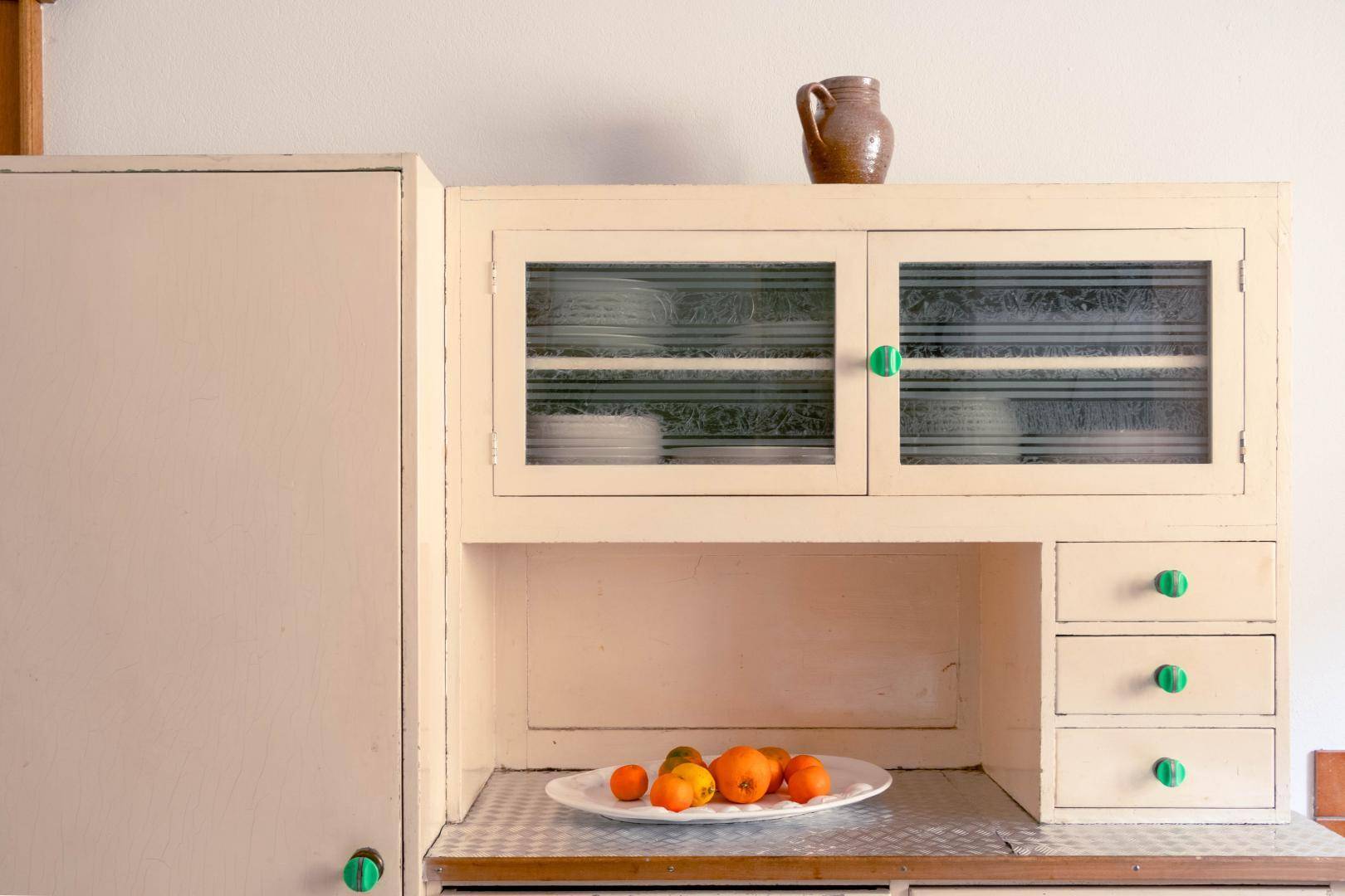 Nostalgischer Küchenschrank mit Glastüren und grünen Knöpfen, gefüllt mit Geschirr und Obst.