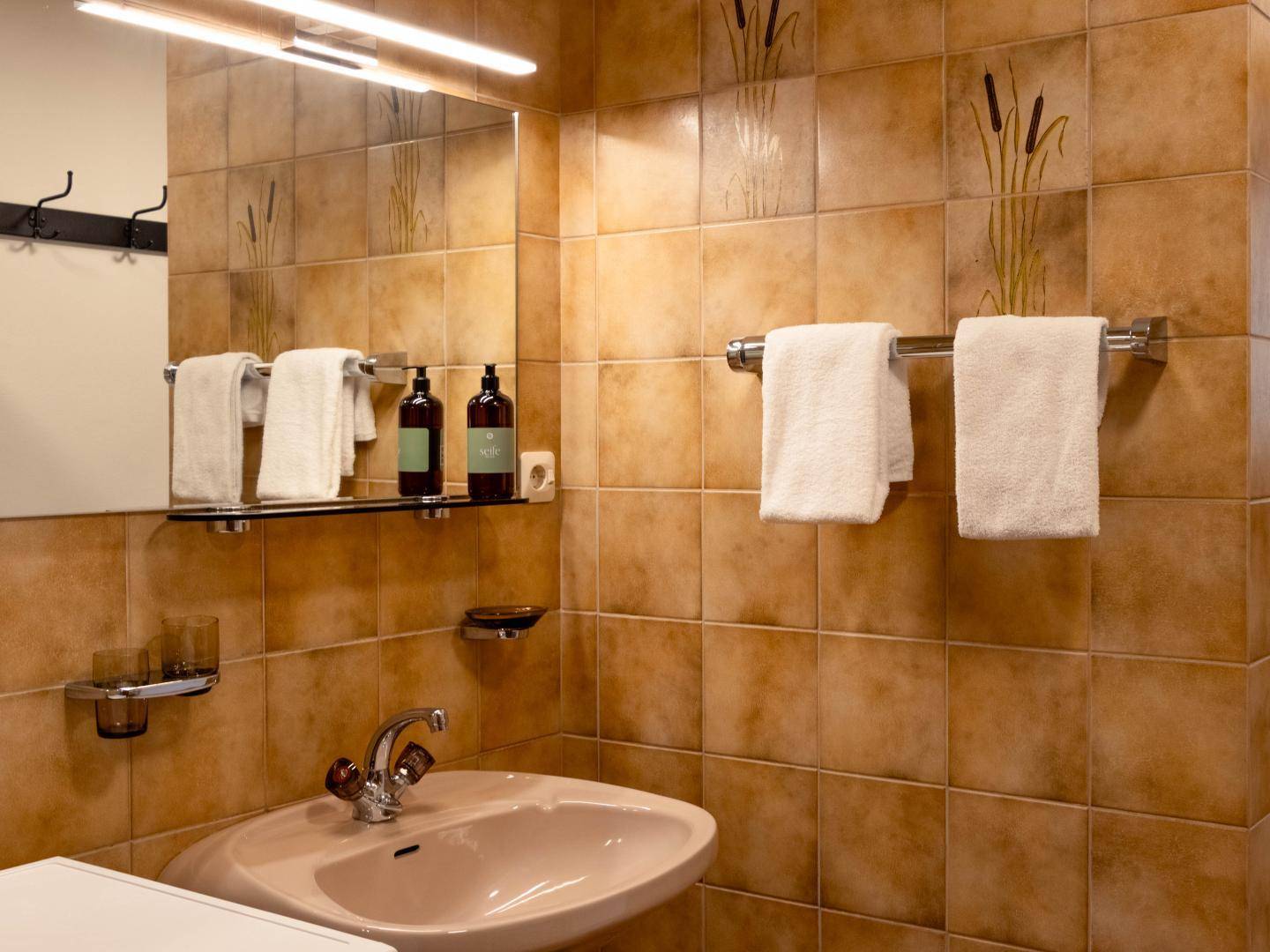 Badezimmer mit braunen Fliesen, Einzelwaschbecken und einem beleuchteten Spiegel.