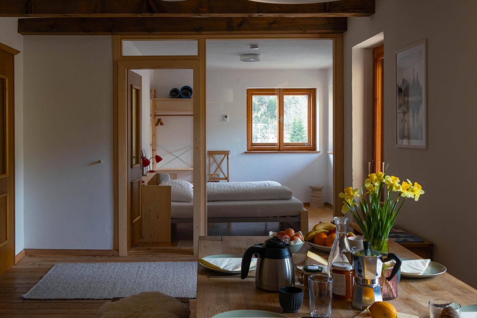 Heller Wohn-Essbereich mit Holzboden und Blick durch eine Glastür in ein Schlafzimmer.