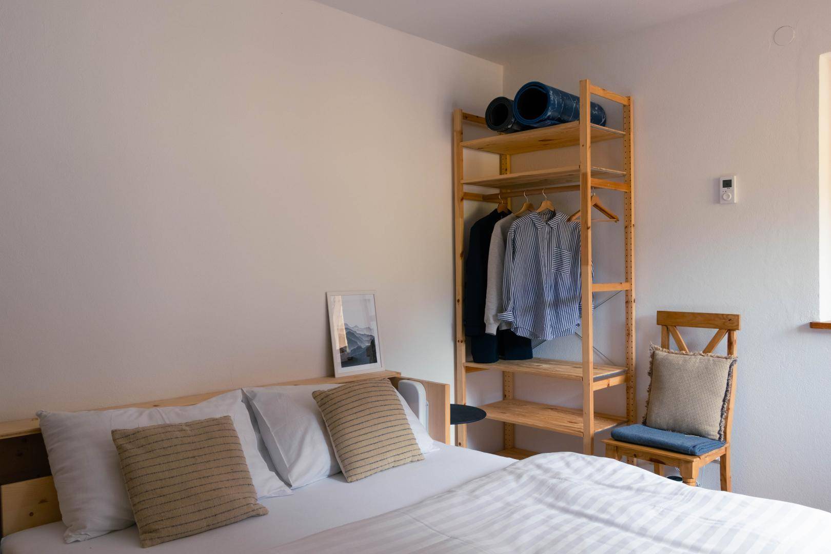 Helles Schlafzimmer mit einfachem Holzbett, offenem Kleiderregal und natürlichem Lichteinfall.