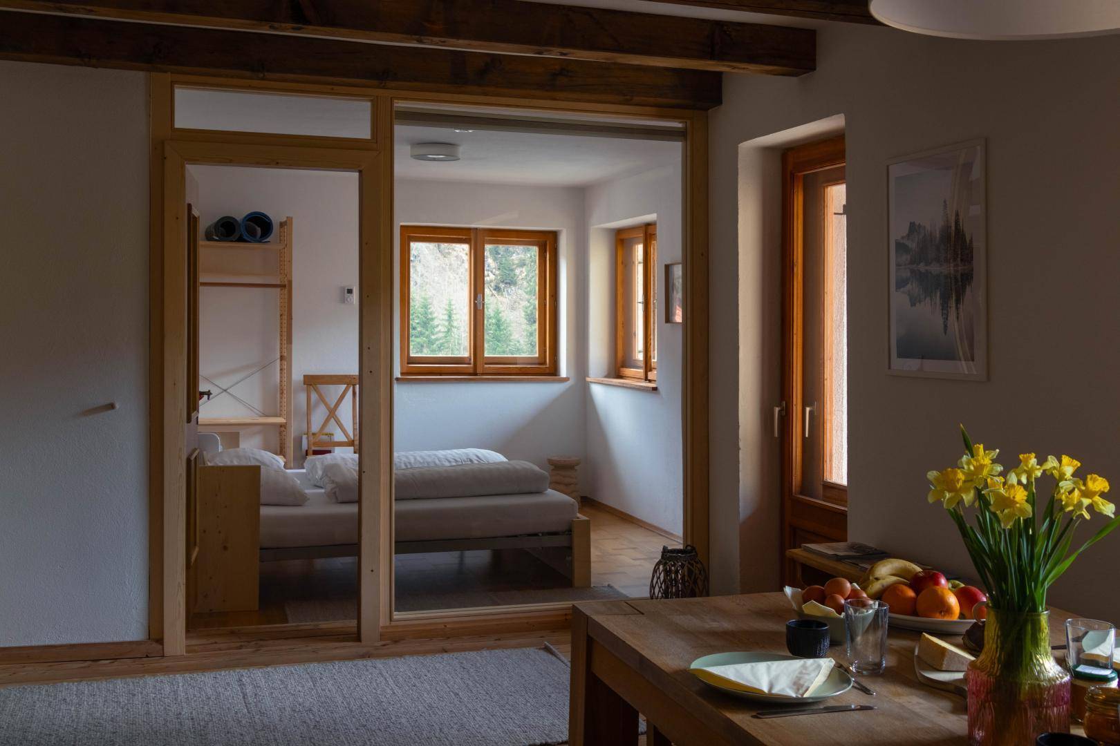 Heller Wohn-Essbereich mit Holzboden und Blick durch eine Glastür in ein Schlafzimmer.