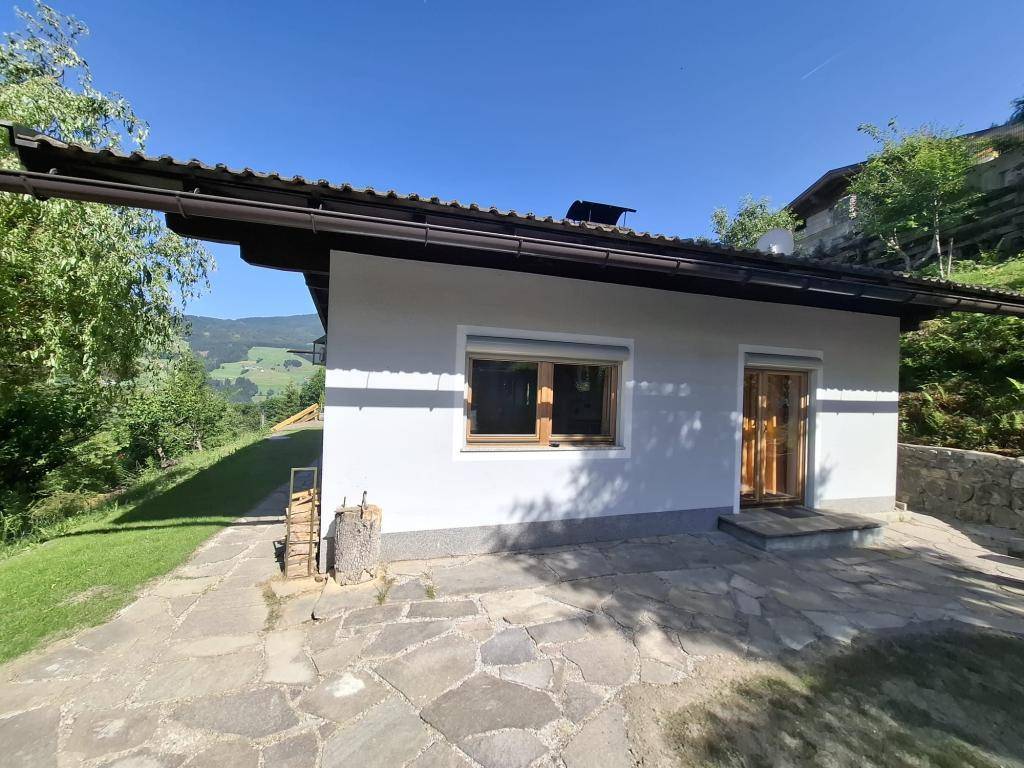 Seitenansicht eines Hauses mit weißer Fassade, Holzfenstern und einem gepflasterten Bereich im Sonnenschein.