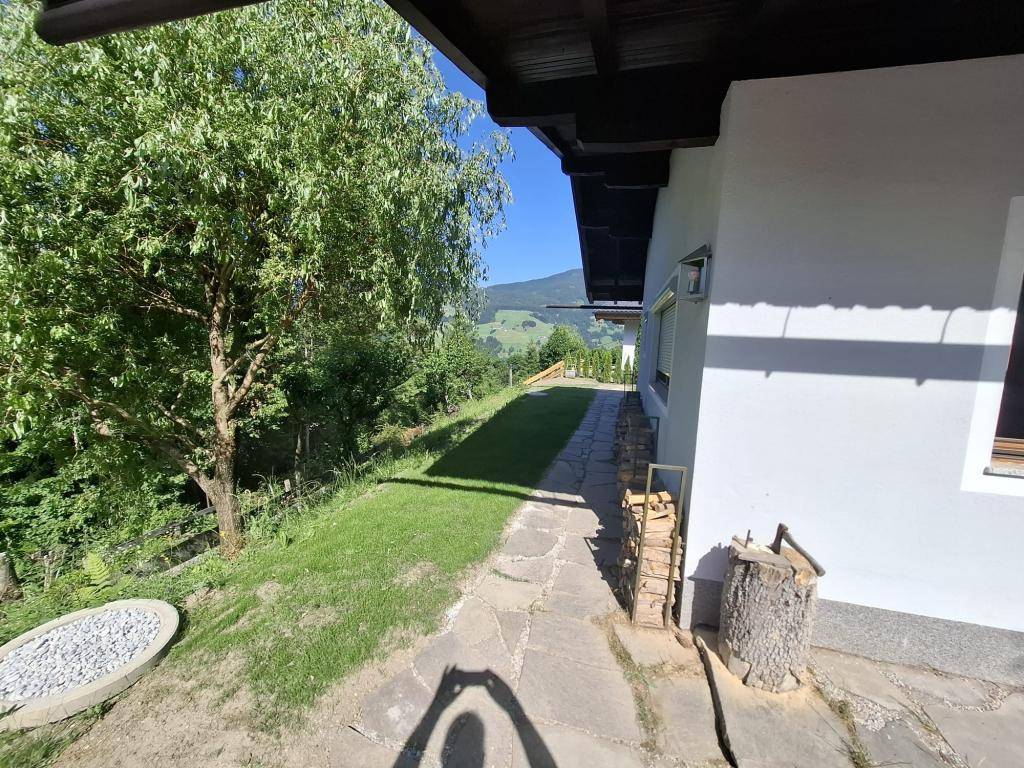 Langer Gartenweg entlang des Hauses mit Holzstapel und weitem Blick auf die grüne Landschaft im Sonnenschein.