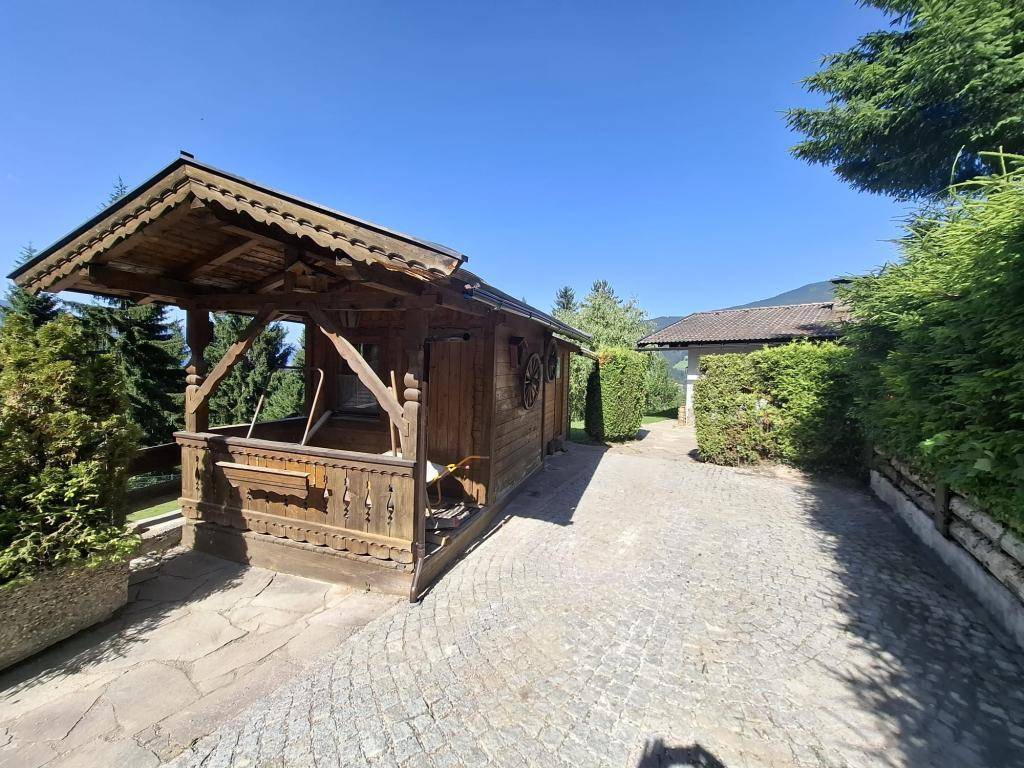 Traditionelles Holzhaus mit Schnitzereien neben der gepflasterten Einfahrt, umgeben von Grün unter blauem Himmel.