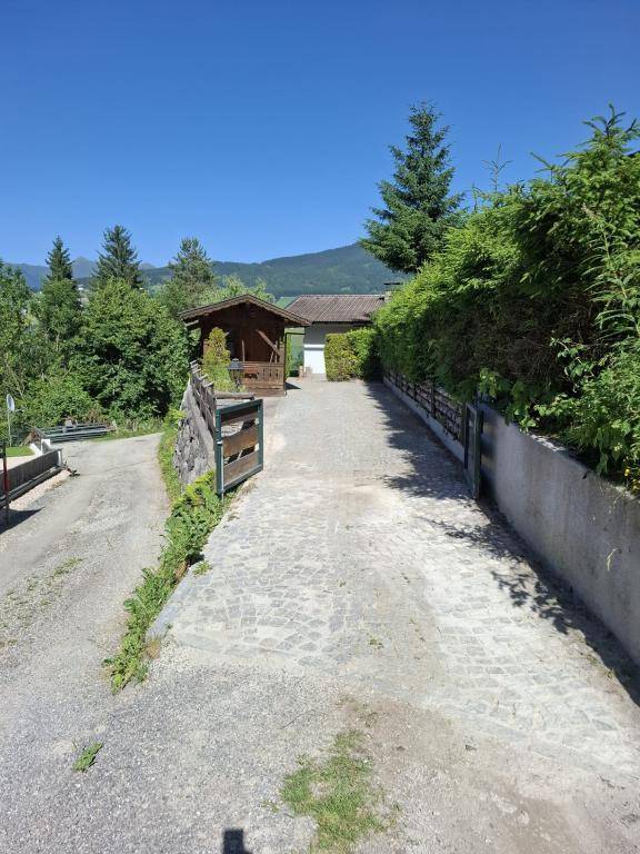 Offene Einfahrt mit Kopfsteinpflaster, umgeben von Hecken und Bäumen, führt zum Haus unter blauem Himmel.