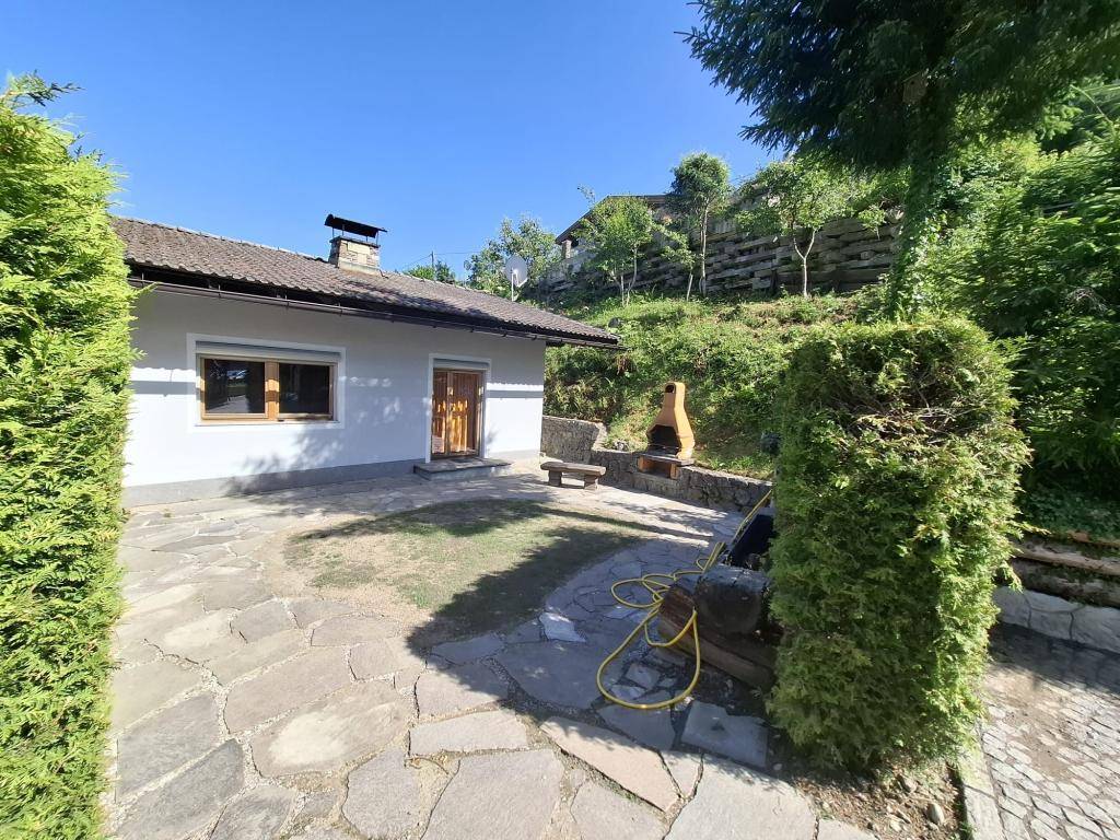 Gepflasterte Terrasse hinter dem Haus mit eingebautem Grill und Blick auf die grüne Umgebung im Sonnenschein.