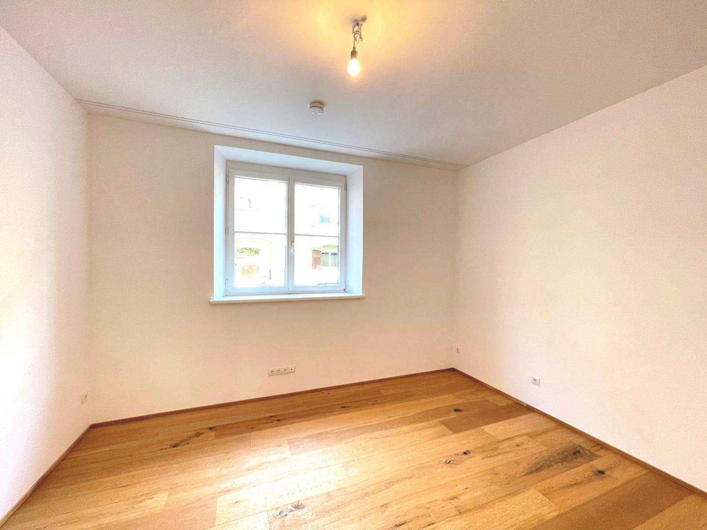 Leerer Raum mit Holzboden, weiß gestrichenen Wänden und einem klassischen Fenster.