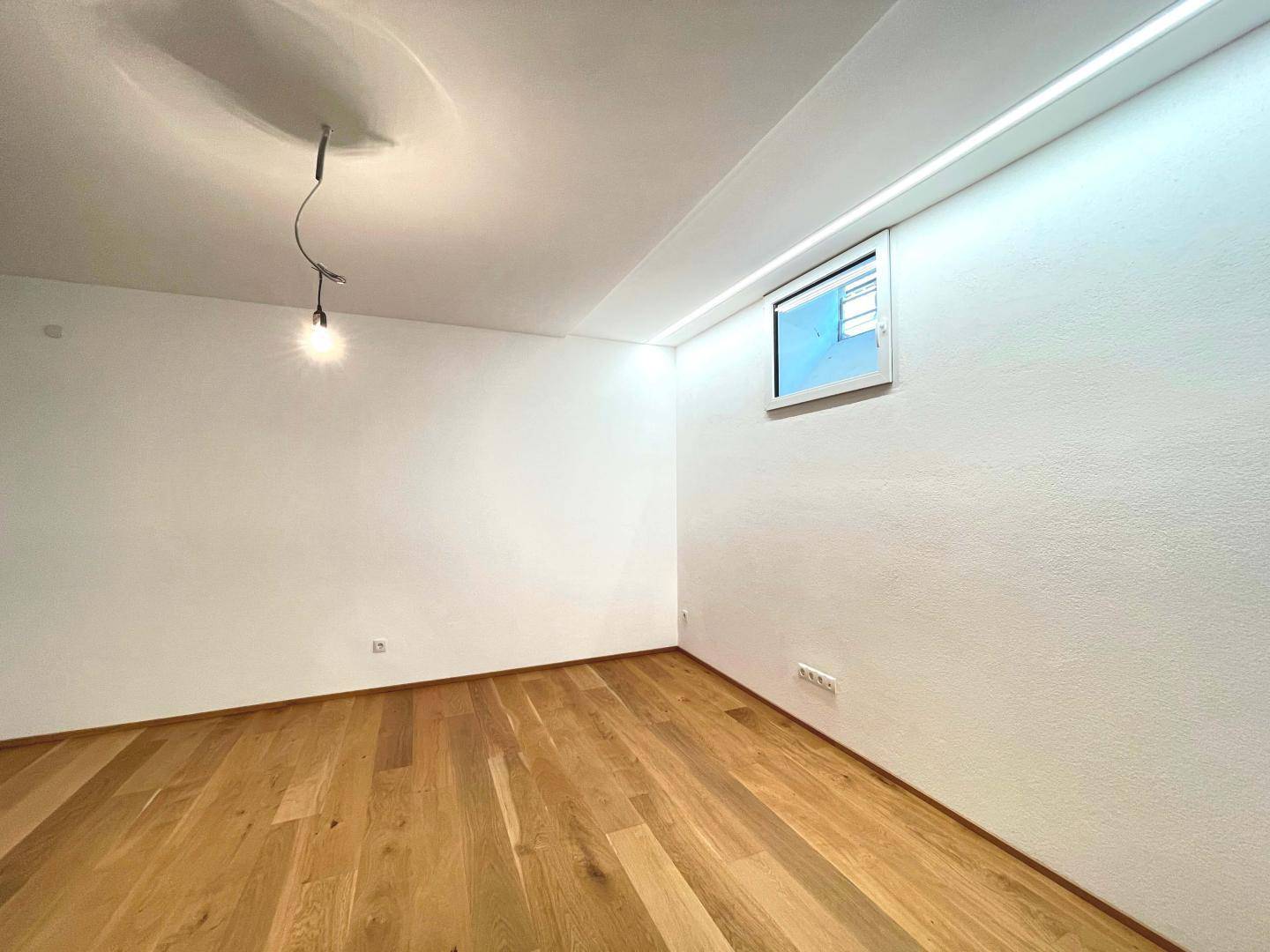Leerer Raum mit Holzboden, weiß gestrichenen Wänden und einem kleinen Fenster.