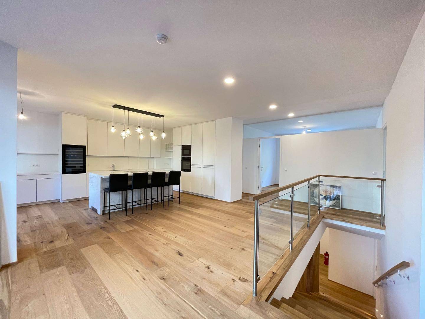 Offener Wohnbereich mit moderner Küche, Holzboden und eleganter Treppe mit Glasgeländer.