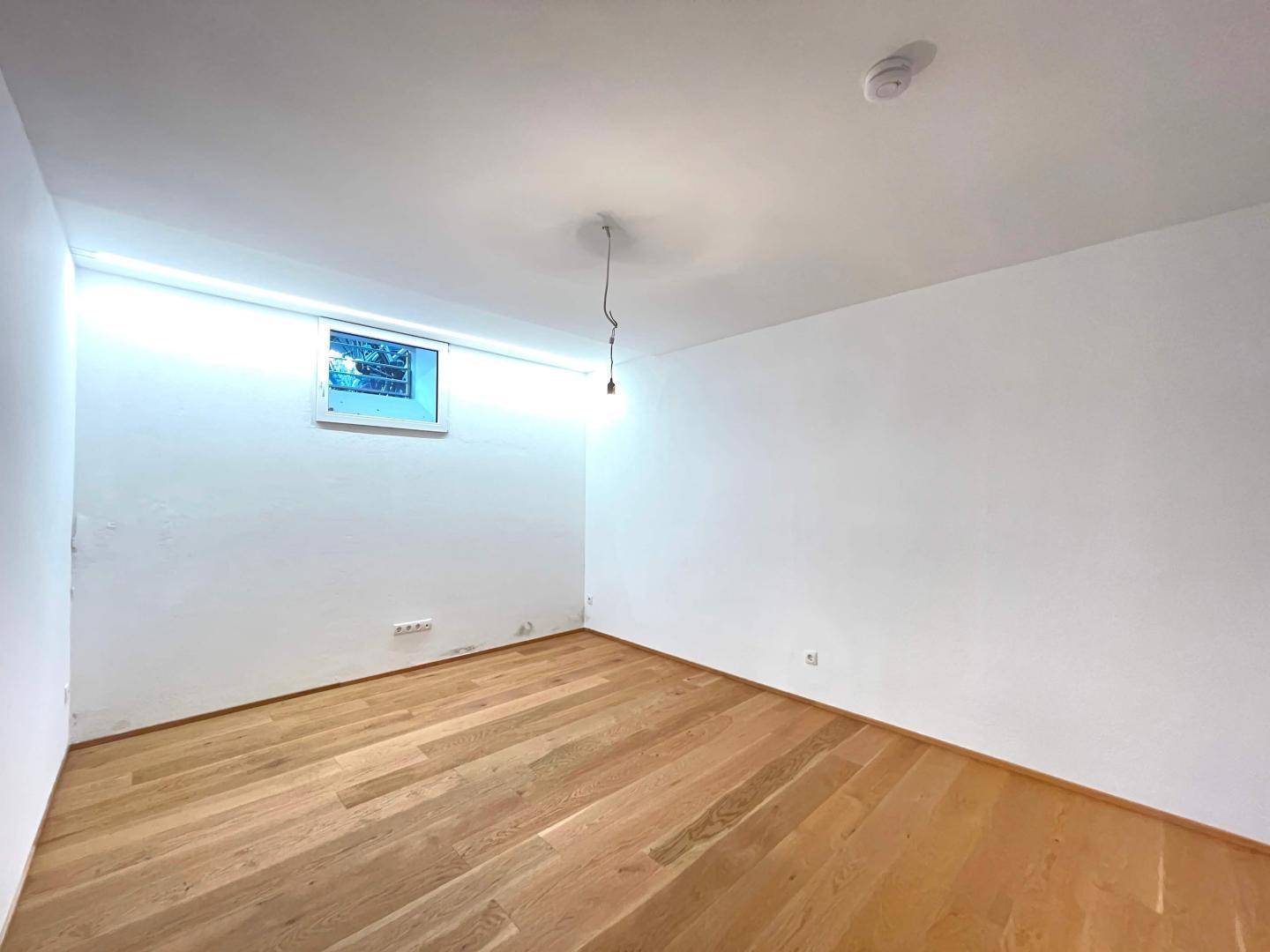 Heller, leerer Raum mit Holzboden, weiß gestrichenen Wänden und einem kleinen Fenster.