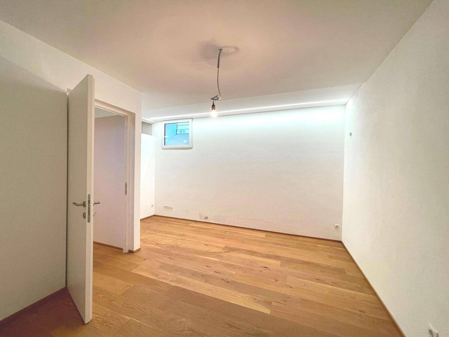 Heller, leerer Raum mit Holzboden, weiß gestrichenen Wänden und einer Tür.