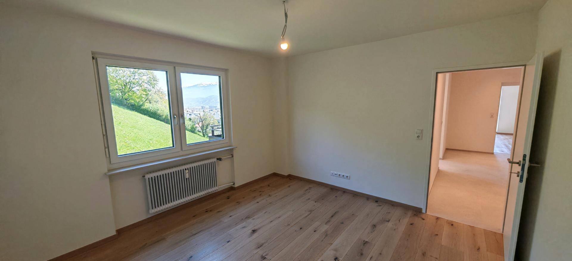 Unmöbliertes Schlafzimmer mit Holzboden, großem Fenster und Blick ins Grüne.