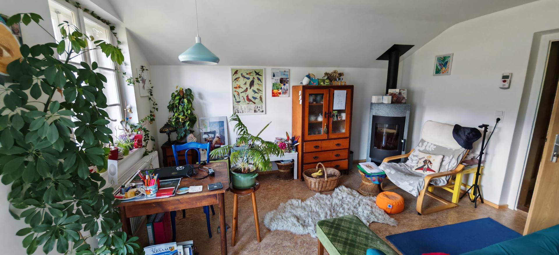 Gemütliches Wohnzimmer mit Holzofen, Schreibtisch und großen Fenstern, die viel Tageslicht hereinlassen.