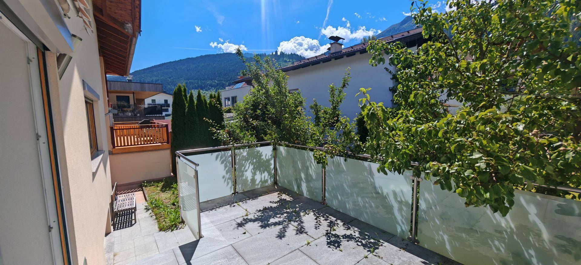 Sonniger Terrassenbereich mit Glasgeländer, umgeben von Bäumen und Bergblick.