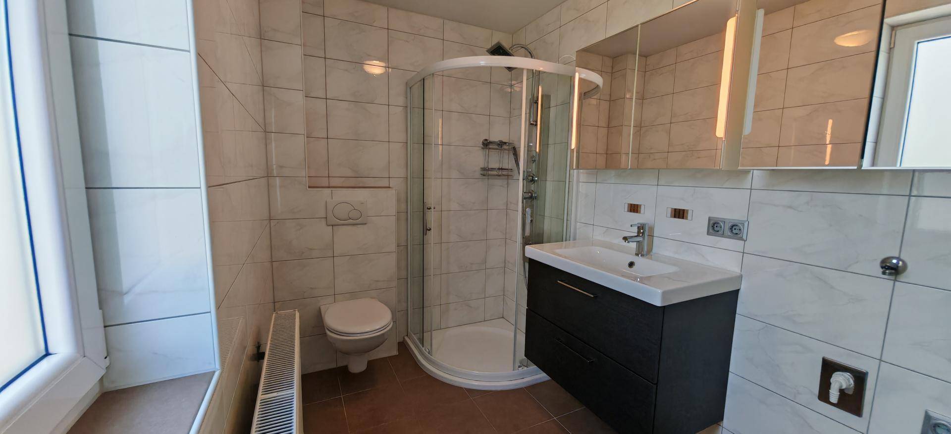 Badezimmer mit Eckdusche, WC, Waschtisch und Fenster, hell gefliest.
