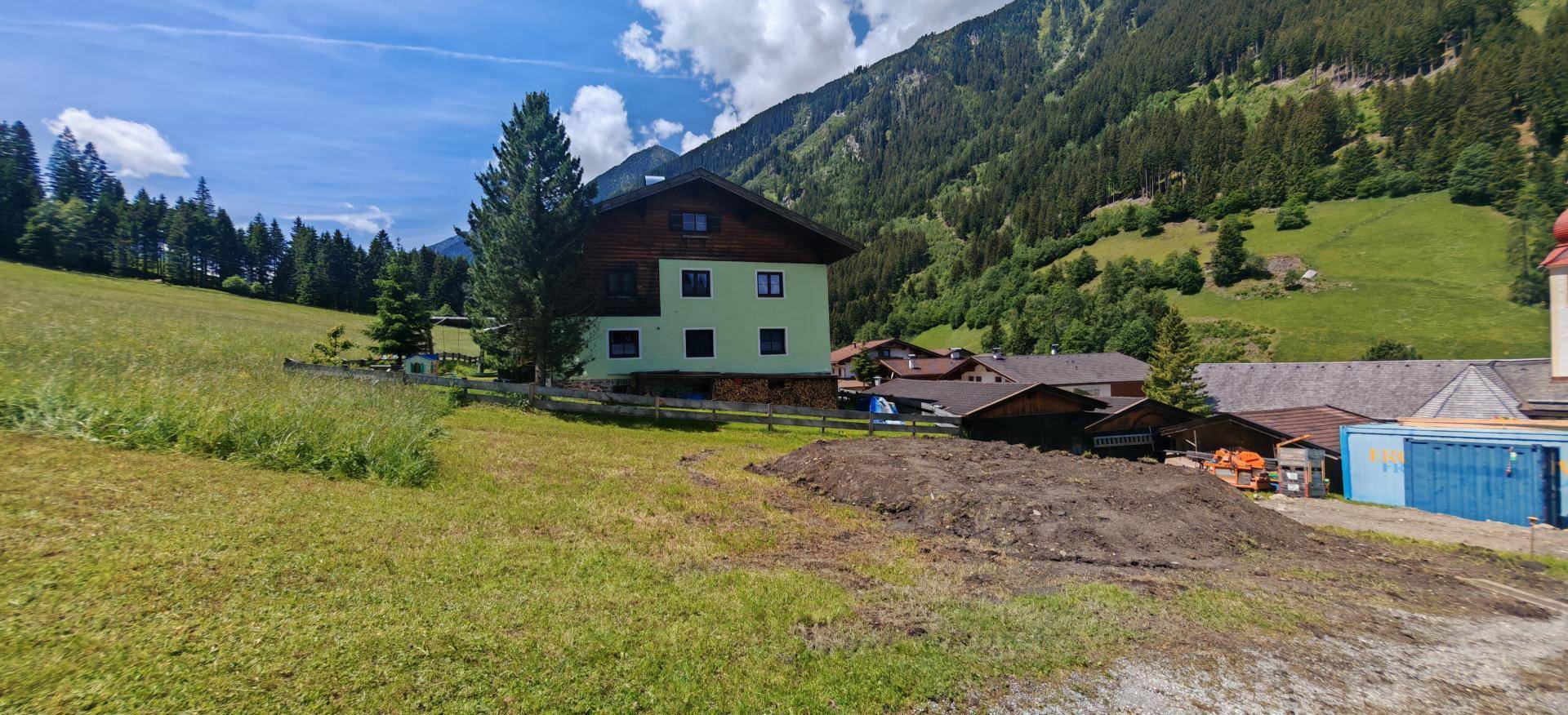 Grüne Wiesen und ein Baugrundstück vor einem traditionellen Haus mit Bergen und Wald im Hintergrund.