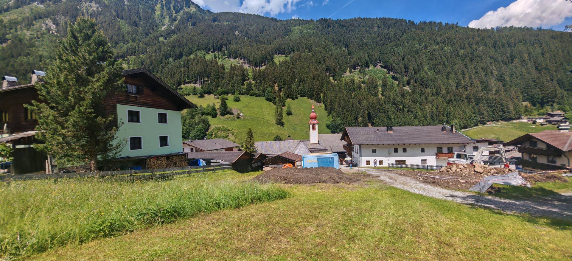 Blick auf Häuser in einem alpinen Dorf mit grünen Wiesen, Wäldern und majestätischen Bergen im Hintergrund.