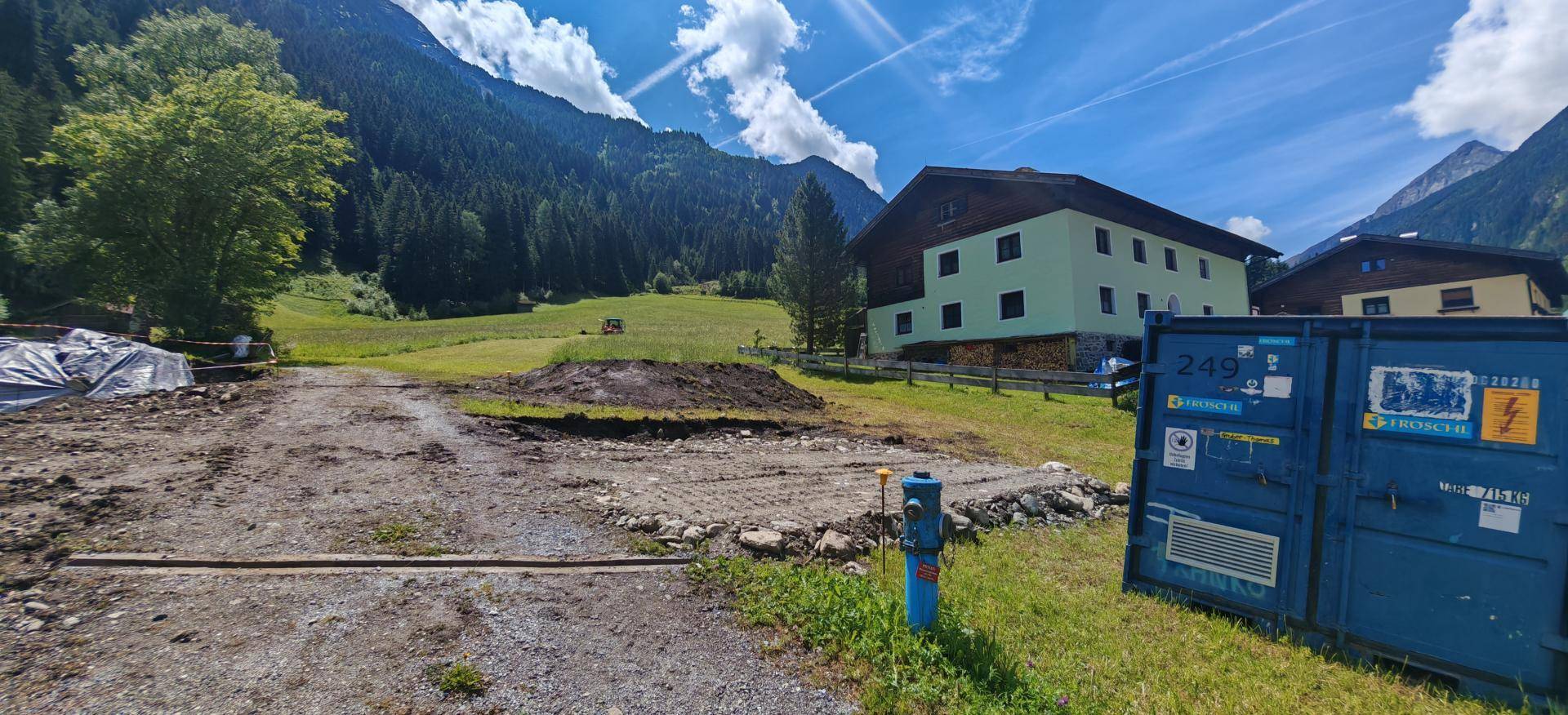 Baugrundstück mit einem blauen Container, umgeben von Häusern und der beeindruckenden Berglandschaft.