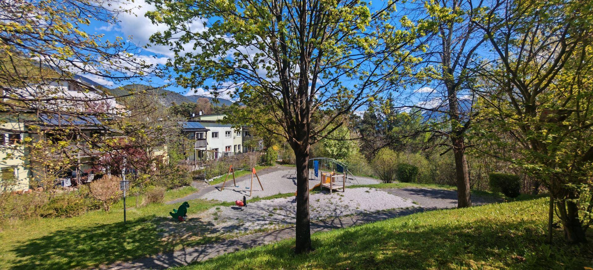 Kinderspielplatz im Freien mit Schaukeln und Rutsche, umgeben von Bäumen.