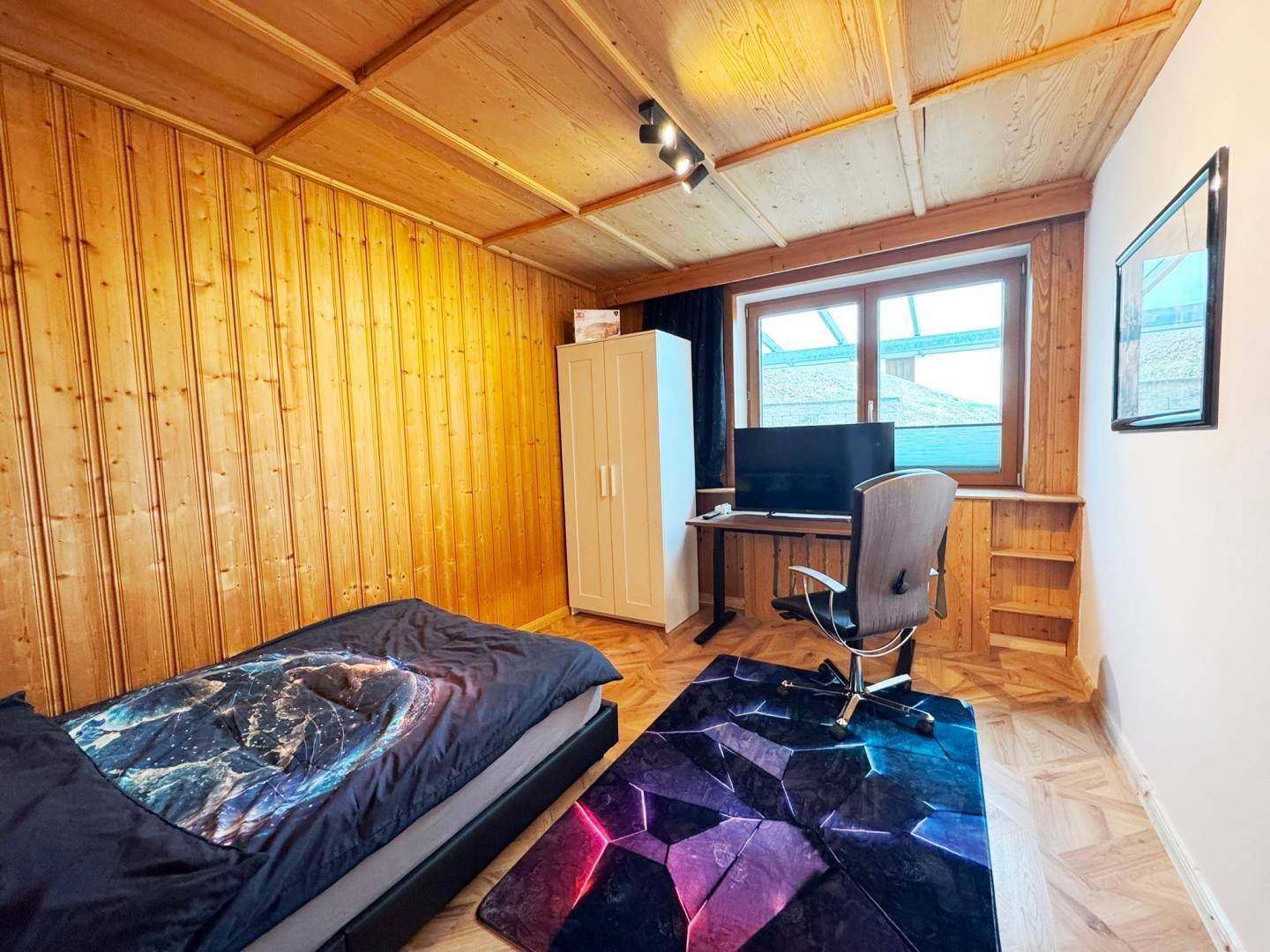 Schlafzimmer mit Holzwänden und -decke, Bett, Schreibtisch und Fenster.